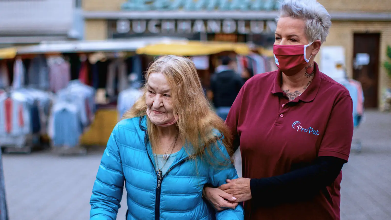 Zwei Frauen in einer Fußgängerzone. Die eine Frau stützt die andere Frau, die einen Rollator benutzt.