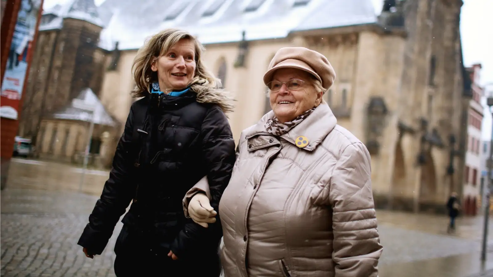 Zwei Frauen in einer Fußgängerzone. Die eine Frau stützt die andere Frau, die einen Rollator benutzt.