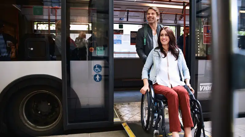 Ein junger Mann schiebt eine junge Frau im Rollstuhl die Rampe eines Busses hinunter