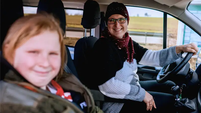 Eine Frau sitzt am Steuer eines Fahrzeugs und schaut gemeinsam mit einer Jugendlichen in die Kamera.