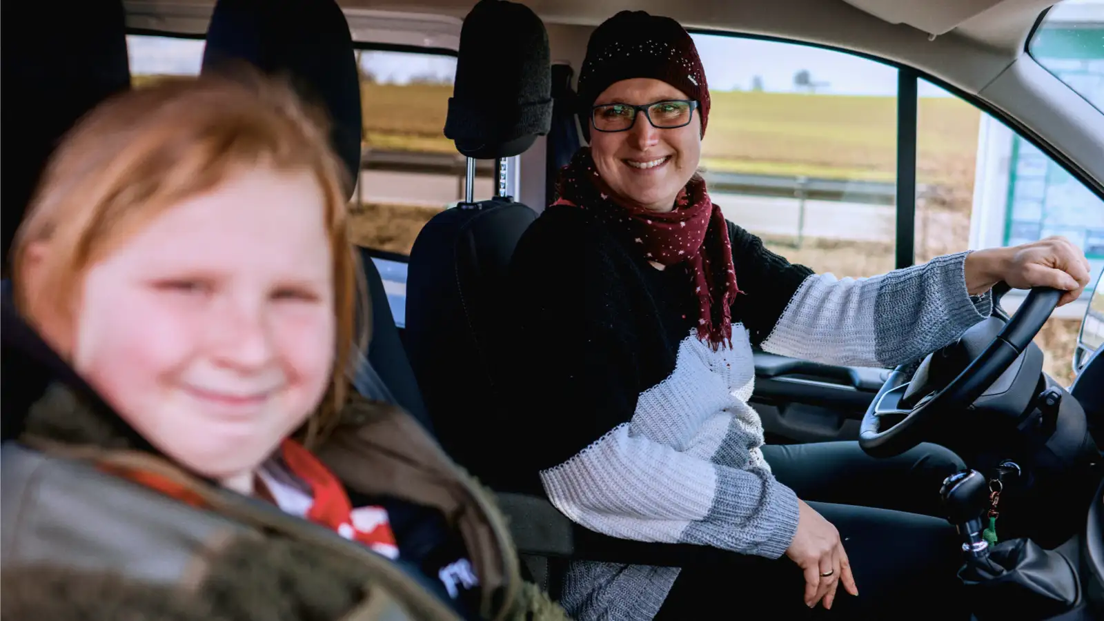 Eine Frau sitzt am Steuer eines Fahrzeugs und schaut gemeinsam mit einer Jugendlichen in die Kamera.
