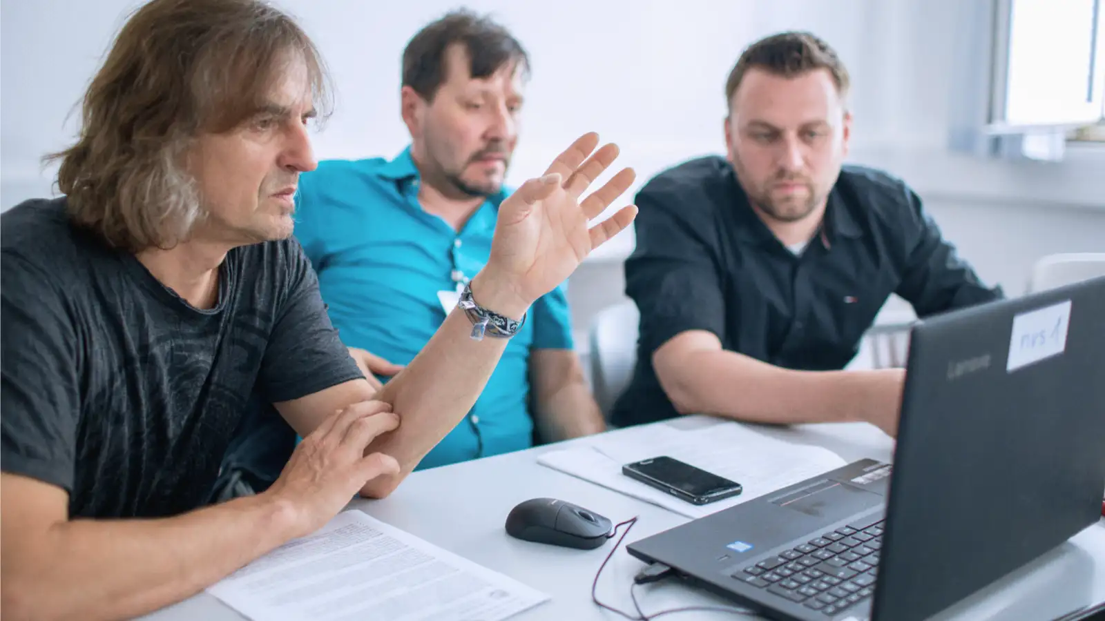 Drei Männer sitzen am Schreibtisch und schauen gemeinsam auf einen Laptop-Bildschirm