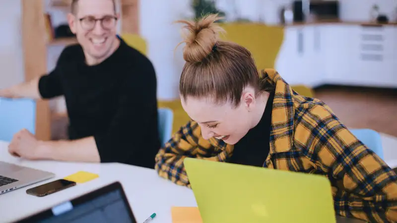Eine junge Frau und ein Mann sitzen lachend vor ihren Laptops