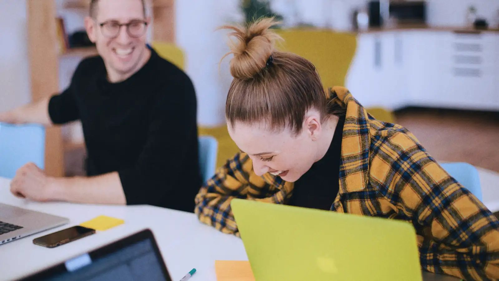 Eine junge Frau und ein Mann sitzen lachend vor ihren Laptops