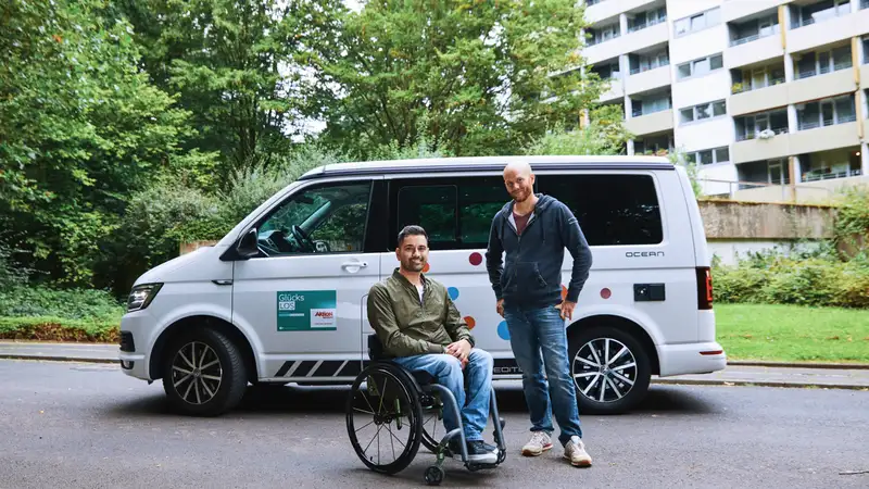 Zwei Männer auf einer Straße vor einem Transporter. Einer der beiden Männer sitzt im Rollstuhl.