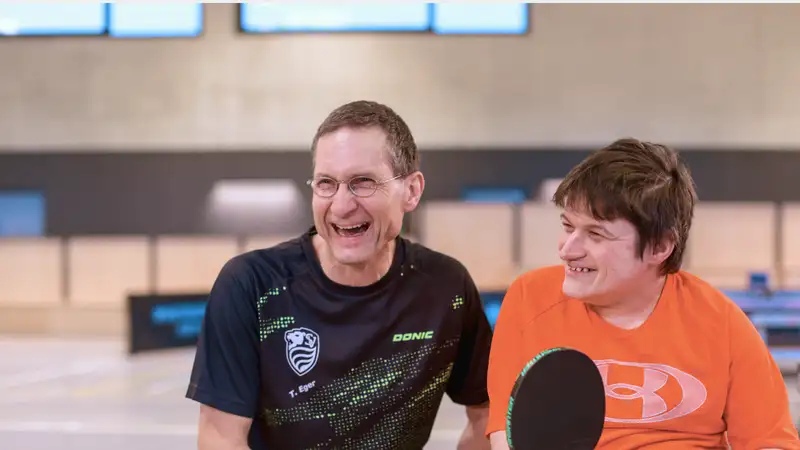 Zwei Personen in einer Turnhalle. Eine davon sitzt im Rollstuhl und hält einen Tischtennisschläger.