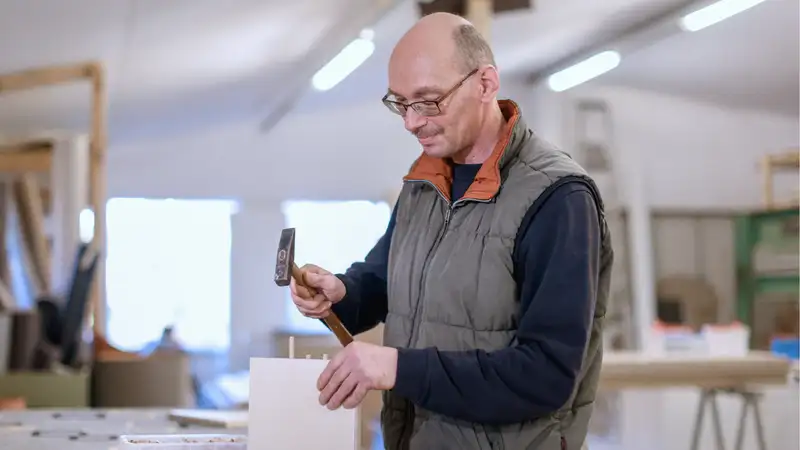 Ein Mann in einer Werkstatt, der mit einem Hammer Zapfen in ein Werkstück schlägt