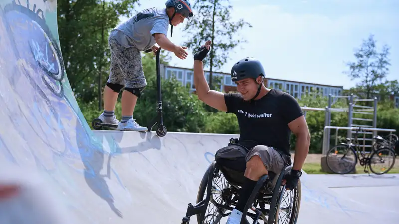 Ein Junge mit einem Scooter und ein Mann im Rollstuhl auf einer Skateranlage