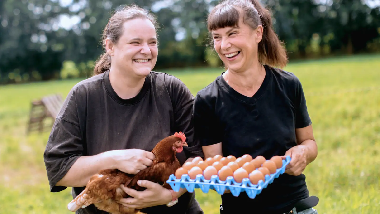 Zwei lachende Frauen halten ein Huhn und eine Schale mit Eiern.