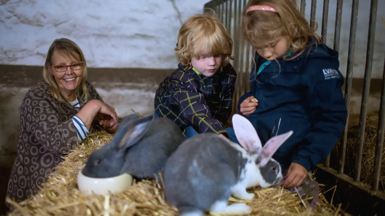 Zwei Kinder und eine Frau in einem Stall mit zwei Kaninchen.