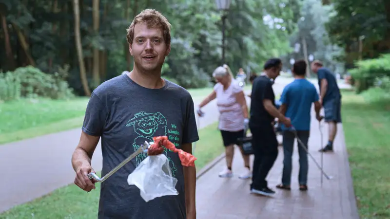 Ein junger Mann beim Müllsammeln im Park, hält mit der Zange eine Plastiktüte.