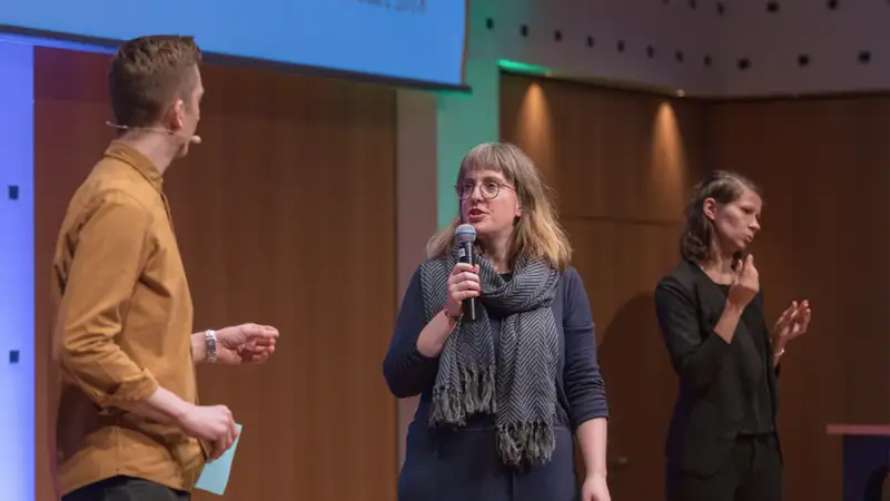 Zwei Personen unterhalten sich auf einer Bühne. Die Unterhaltung wird von einer Dolmetscherin gebärdet.