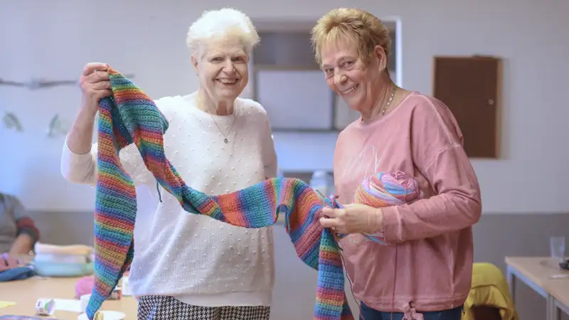 Zwei lachende ältere Damen zeigen einen bunten gestrickten Schal.