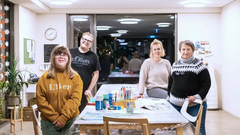 Vier Menschen an einem Tisch mit Pinseln und Farben schauen in die Kamera.