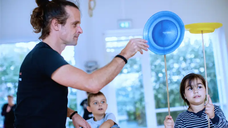 Kinder jonglieren unter Anleitung eines Mannes mit Tellern auf Holzstäben.