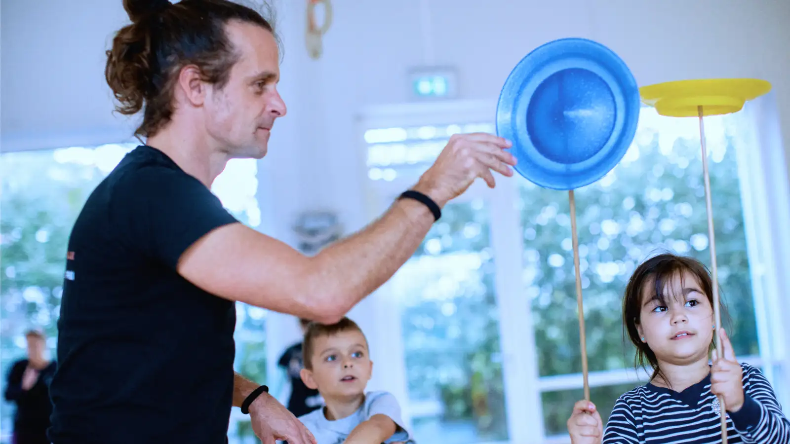 Kinder jonglieren unter Anleitung eines Mannes mit Tellern auf Holzstäben.