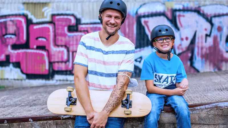 Ein Kind und ein Mann sitzen vor einer Graffiti besprühten Mauer. Der Mann hält ein Skateboard in der Hand.