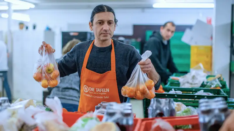 Mitarbeiter einer Tafel vor verschiedenen Kisten mit Obst und Gemüse. Er hält zwei gefüllte Plastiktüten mit Obst in der Hand.