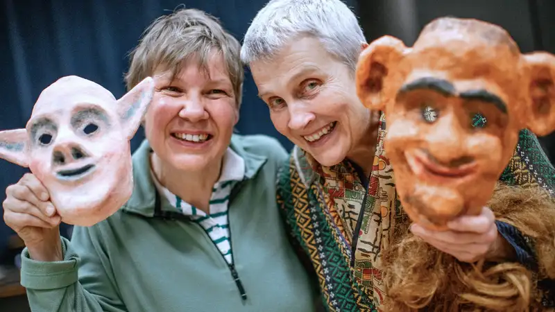 Zwei lachende Frauen, die jeweils eine groteske Maske in der Hand halten.