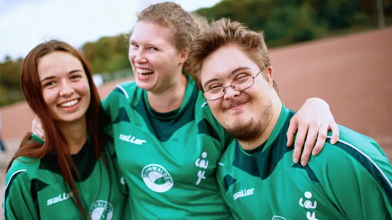 Drei Jugendliche in Sporttrikots umarmen sich und lachen in die Kamera