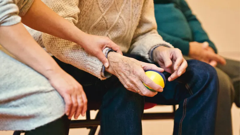 Eine sitzende ältere Person hält einen bunten Ball in der Hand. Eine andere Person fasst sie am Arm.