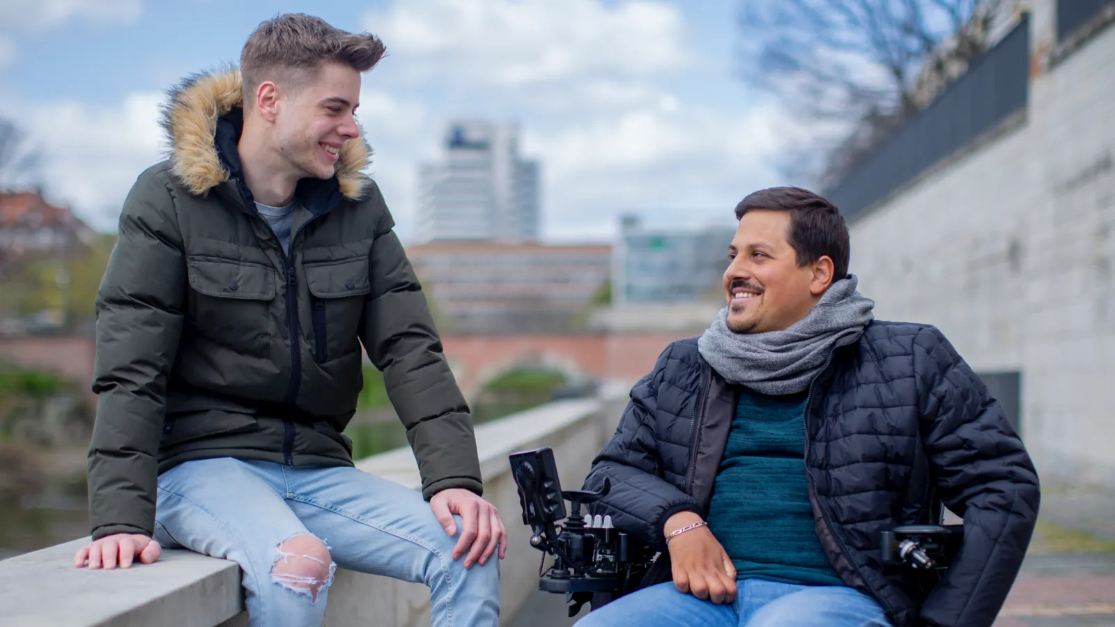 Zwei junge Männer unterhalten sich im Freien. Einer sitzt auf einer Mauer, der andere im Rollstuhl.