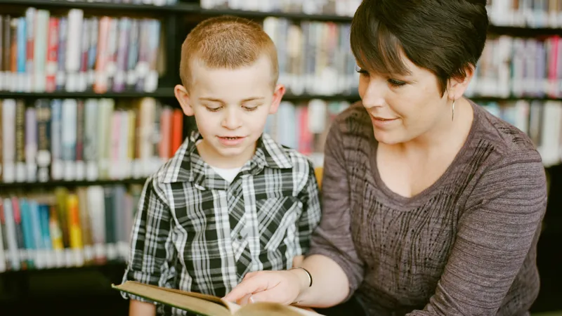 Eine Frau in einer Bibliothek hält ein geöffnetes Buch und liest einem Jungen etwas vor.