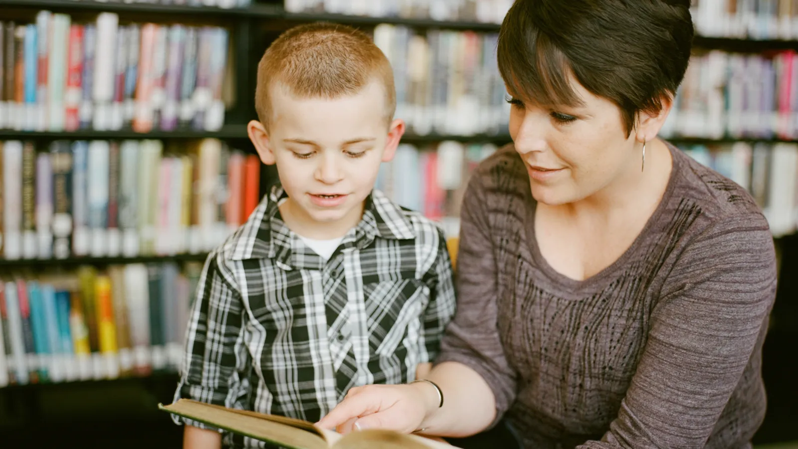 Eine Frau in einer Bibliothek hält ein geöffnetes Buch und liest einem Jungen etwas vor.