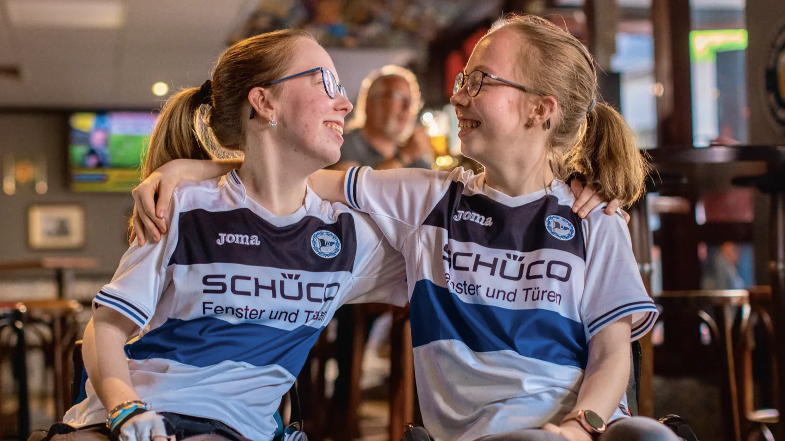 Zwei Mädchen im Rollstuhl mit Fantrikots umarmen sich in einem Café
