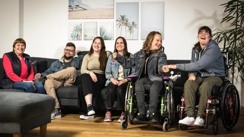 Eine Gruppe von sechs Menschen mit und ohne Behinderung sitzen in einem Wohnzimmer und lachen in Richtung Kamera.