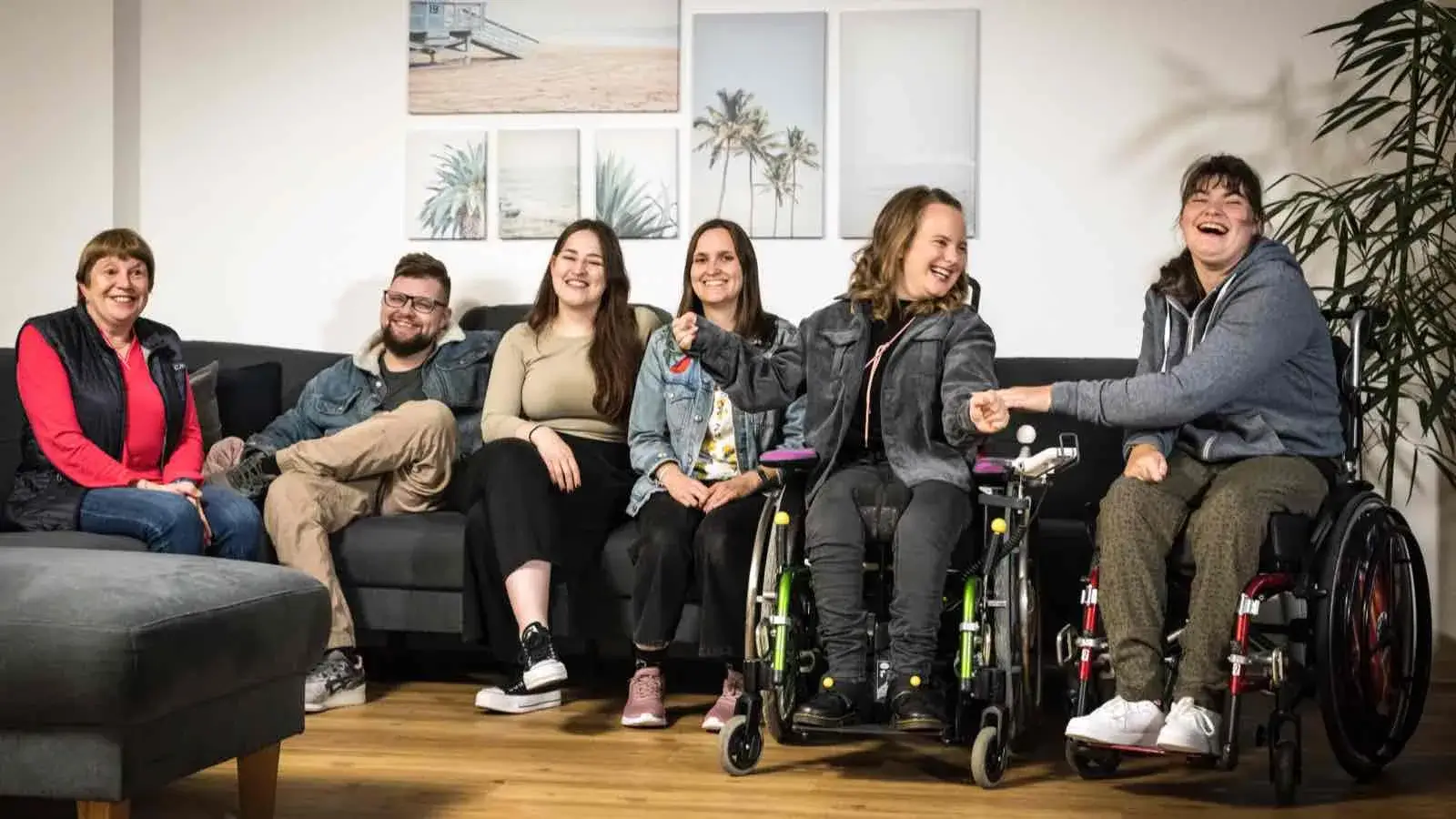 Eine Gruppe von sechs Menschen mit und ohne Behinderung sitzen in einem Wohnzimmer und lachen in Richtung Kamera.