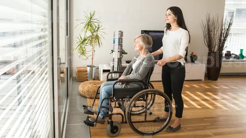 Eine junge Frau schiebt eine Frau im Rollstuhl an ein großes Fenster, aus dem sie hinausblicken