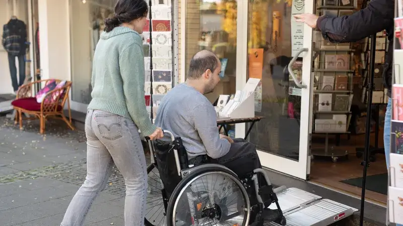 Eine junge Frau schiebt einen Mann im Rollstuhl über eine kleine Rampe in einen Schreibwarenladen.