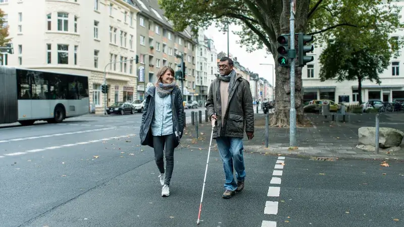 Eine Frau und ein Mann mit Blindenstock überqueren zu Fuß eine Straße mit grüner Ampel.
