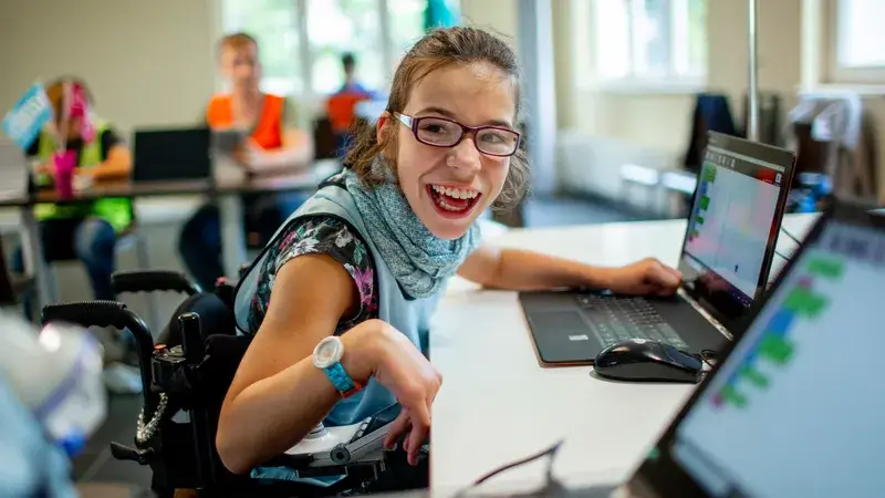 Ein Mädchen mit Behinderung sitzt an einem Computer in einem Gruppenraum und lacht eine andere Person an.