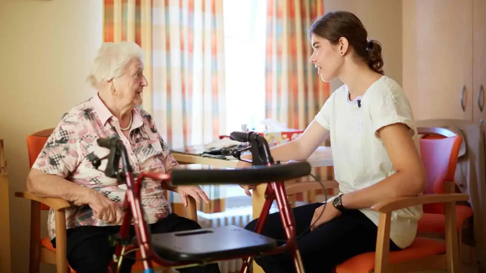 Eine junge Frau sitzt mit einer Seniorin an einem Tisch und unterhält sich mit ihr