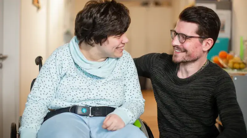 Zwei Männer, einer davon im Rollstuhl, blicken sich lächelnd an.