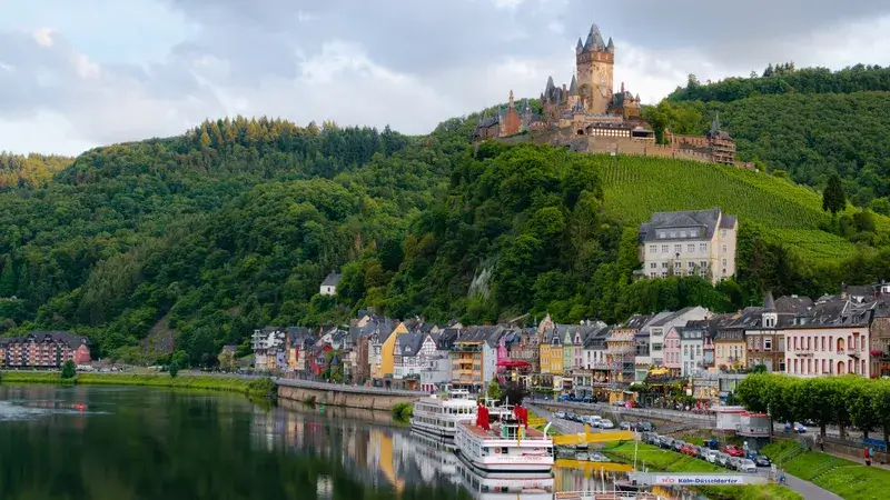 Ein Panorama der Stadt Cochem mit Schiffen auf der Mosel und einer Burg auf dem Berg im Hintergrund.