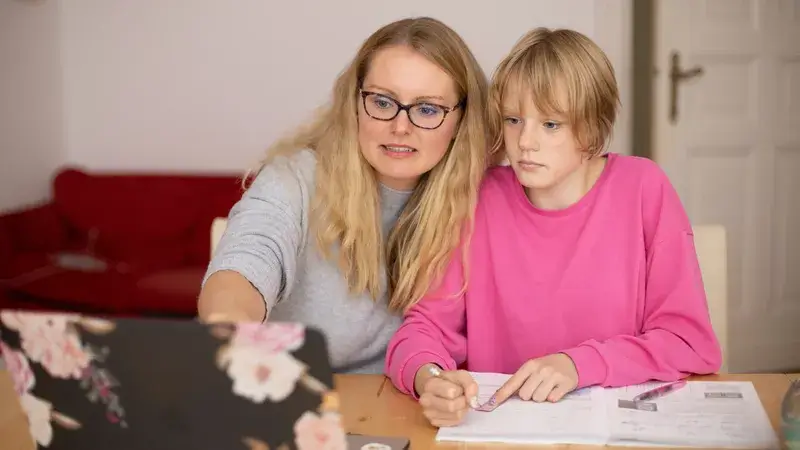 Eine Frau und ein jugendliches Mädchen sitzen an einem Schreibtisch. Die Frau zeigt auf den Laptop-Monitor.