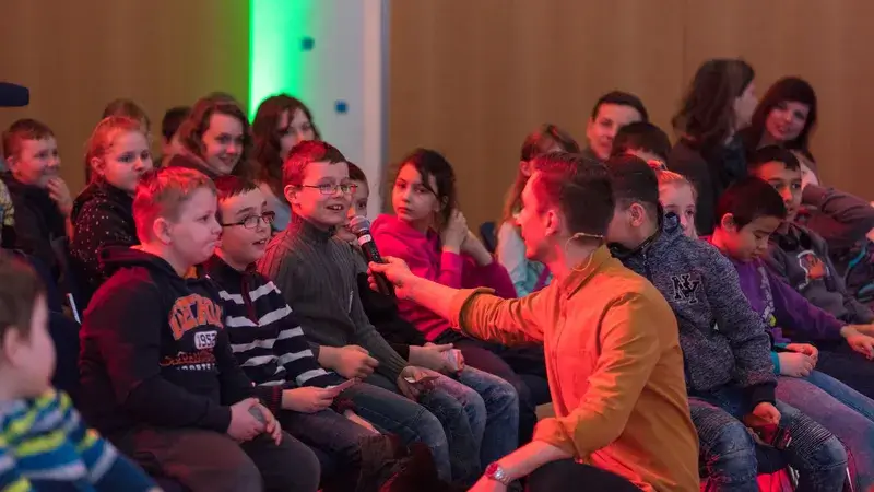 Ein Moderator geht zu einem Publikum aus Kindern und hält einem Kind ein Mikrofon vor das Gesicht