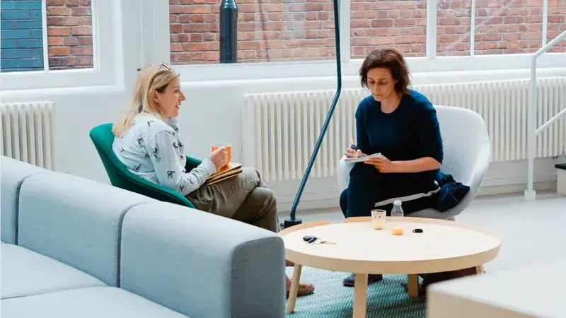 Zwei Frauen sitzen auf Stühlen an einem kleinen runden Tisch und unterhalten sich.