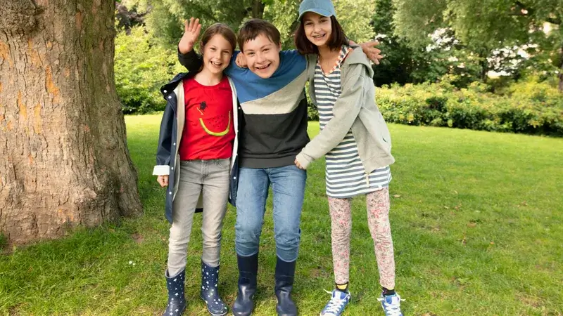 Zwei Mädchen und ein Junge stehen sich umarmend unter einem Baum und lachen.
