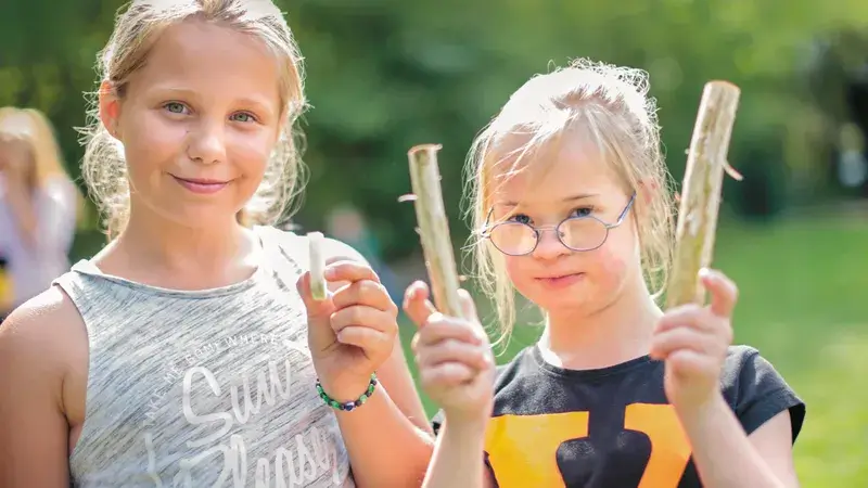 Zwei Mädchen im Freien lächeln in die Kamera; eines hält zwei kurze Stöcke in der Hand.