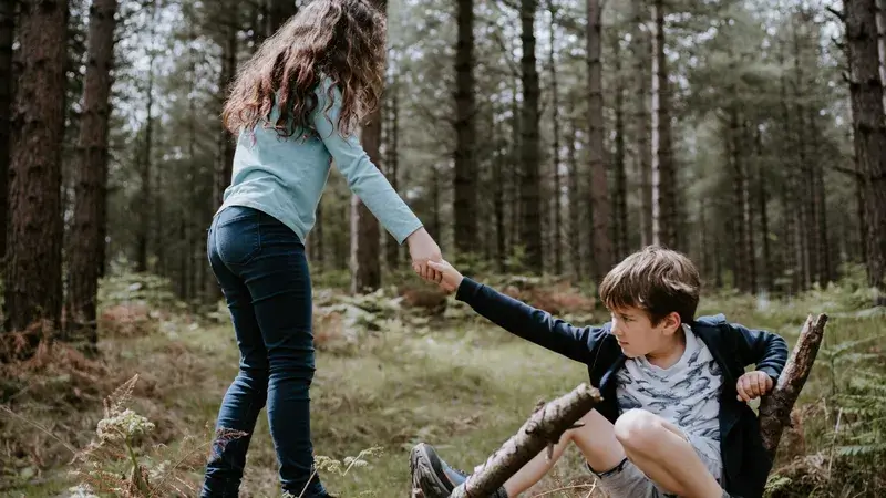 Ein Mädchen hilft einem in der Hocke sitzenden Jungen im Wald auf die Beine.