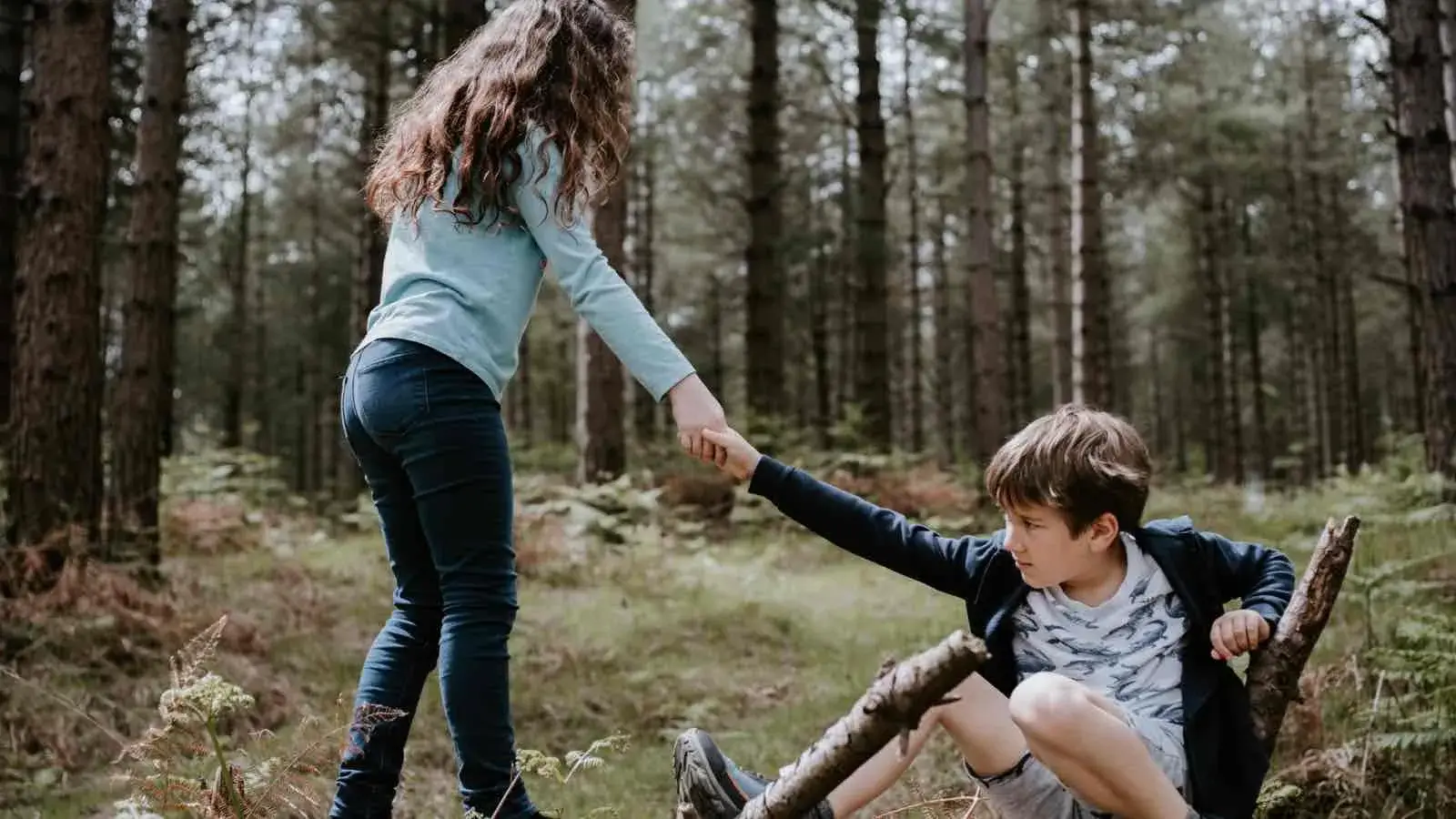 Ein Mädchen hilft einem in der Hocke sitzenden Jungen im Wald auf die Beine.