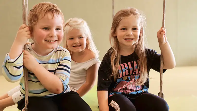 Drei lachende Kinder sitzen gemeinsam auf einer Schaukel in einem Spielzimmer.