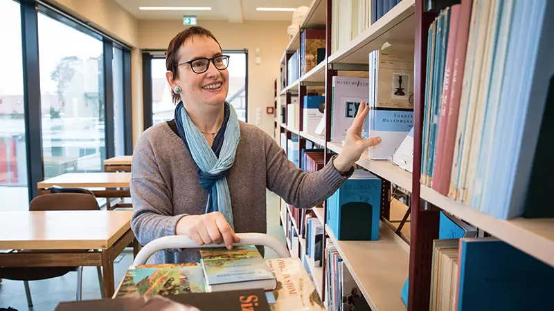 Eine Frau steht in einer Bibliothek zwischen Bücherregalen und sucht nach einem Buch.