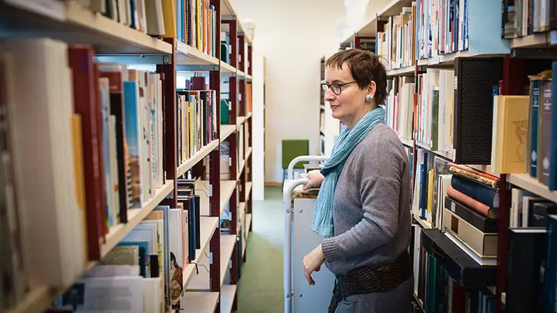 Eine Frau in einer Bibliothek stellt Bücher in ein Regal, während sie neben einem Bücherwagen steht.