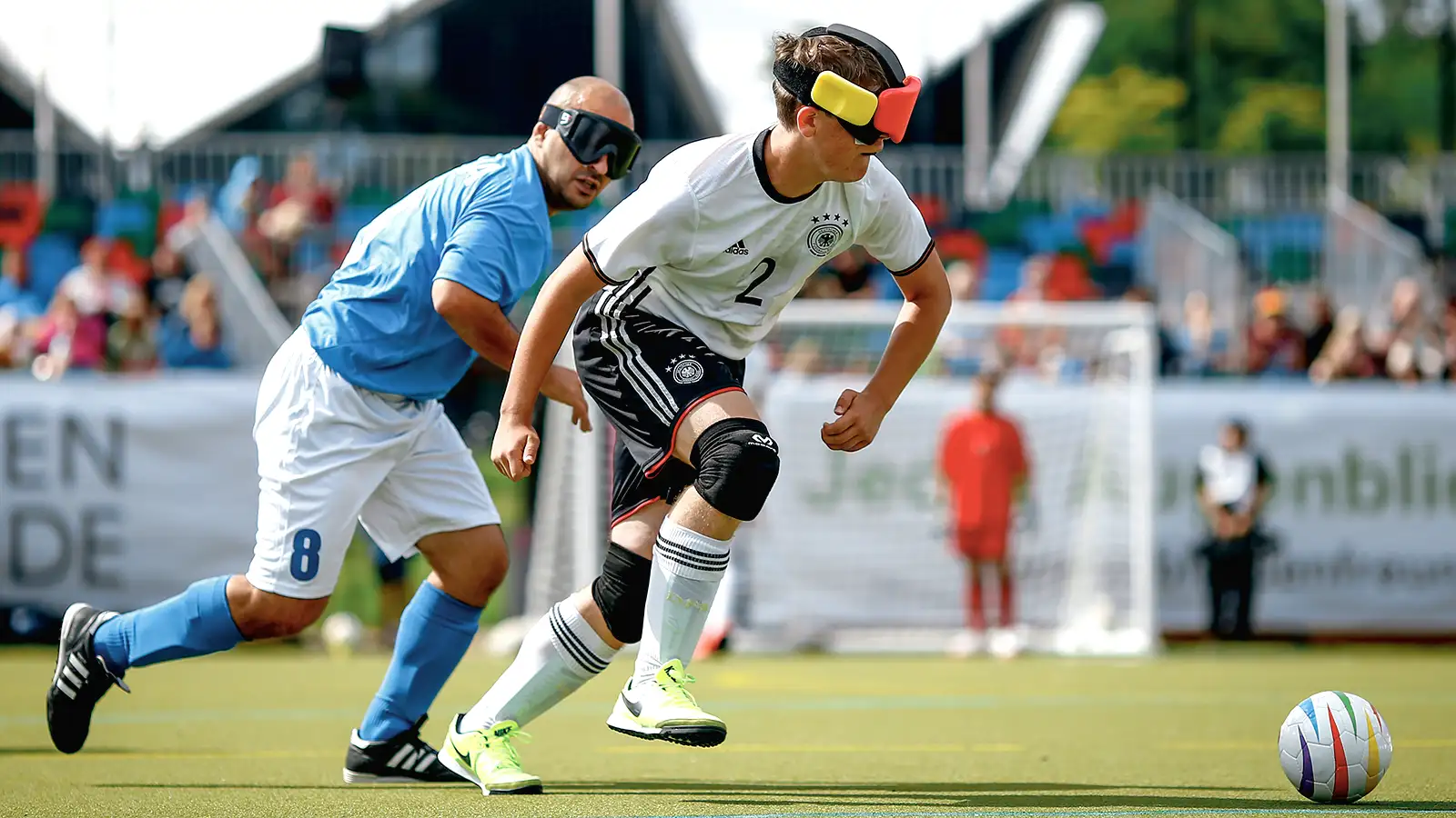 Zwei Blindenfußballspieler, einer in weiß und der andere in blau, kämpfen auf dem Spielfeld um den Ball, beide tragen Augenbinden.