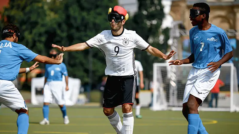 3 Blindenfußballspieler, 2 in blauen Trikots und einer in weiß, auf dem Spielfeld. Der Spieler in weiß kontrolliert den Ball, alle tragen Augenbinden.